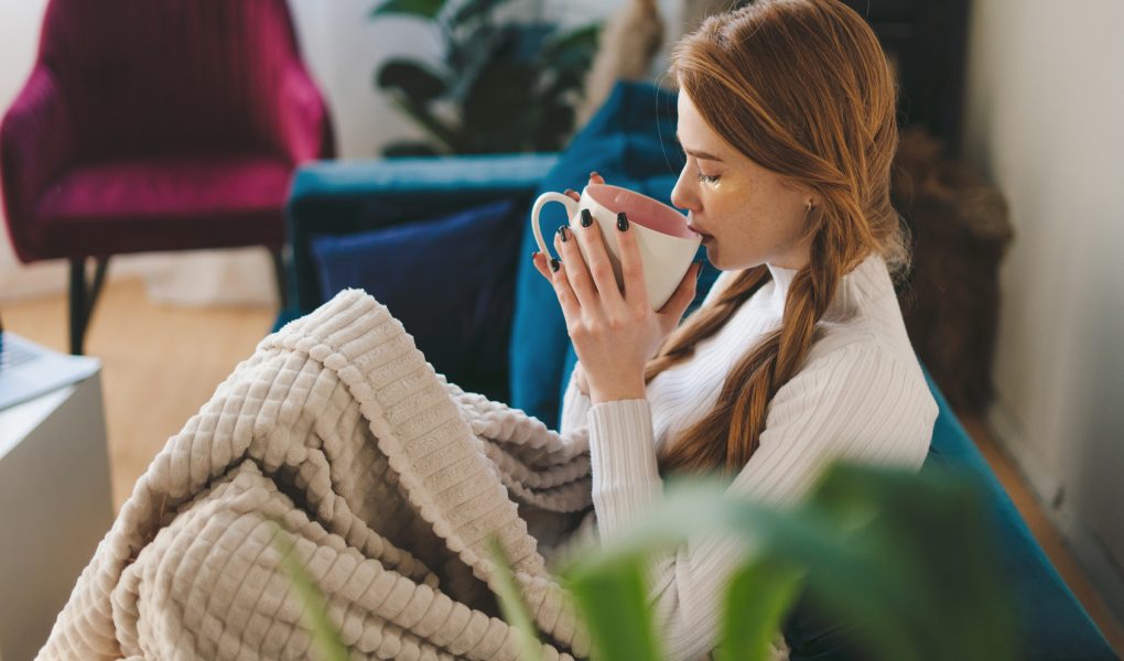 Femme enveloppée dans un plaid sur son canapé, une tasse de thé à la main : se préparer à l'heure d'hiver en douceur