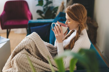 Femme enveloppée dans un plaid sur son canapé, une tasse de thé à la main : se préparer à l'heure d'hiver en douceur